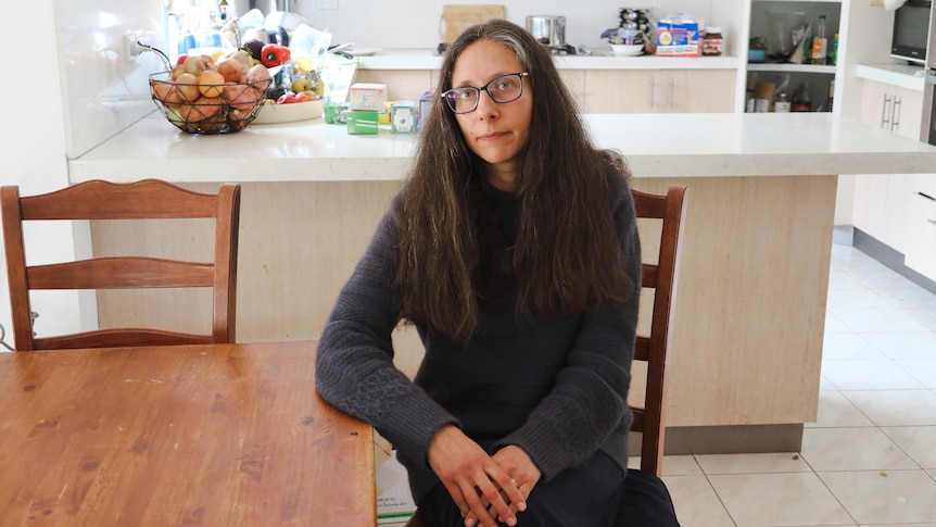 A woman sitting at a dining table looking at the camera with a neutral expression on her face. Behind her is a kitchen.