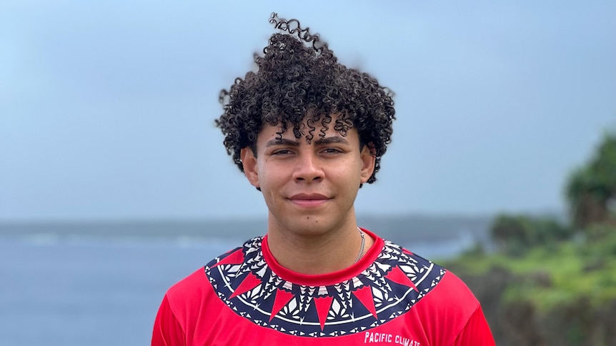 Young man stand in front of coastline, wearing red top and ta'ovala.
