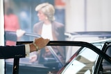 A man's arm rests on a car door. In the background, a photo of Diana walking in a brown blazer is displayed. 