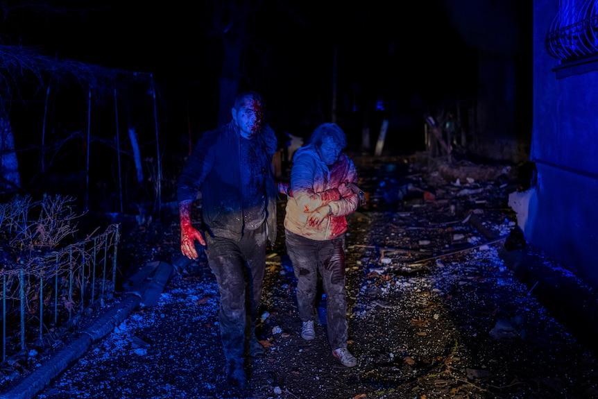 Two people walk in the dark covered in blood and visibly hurt. 