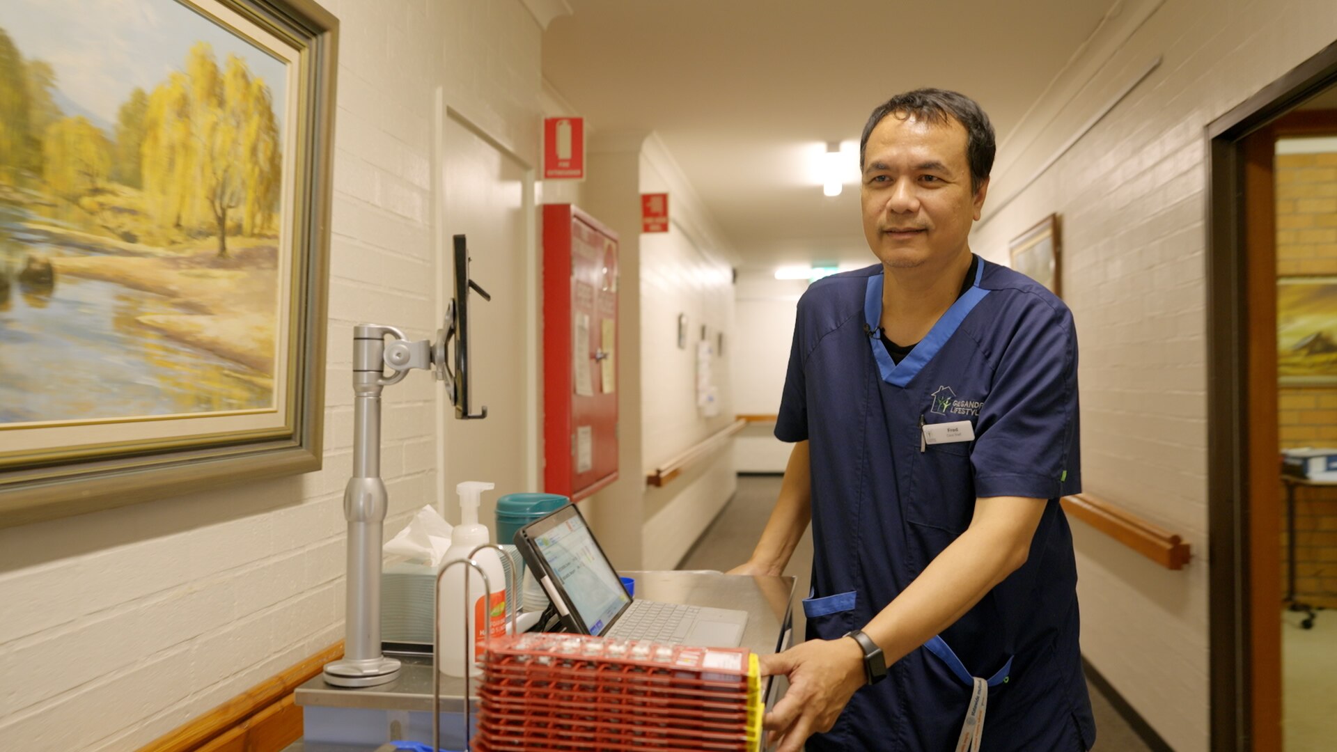 Man dressed in healthcare worker uniform pushes medication trolly down corridor in aged care facility