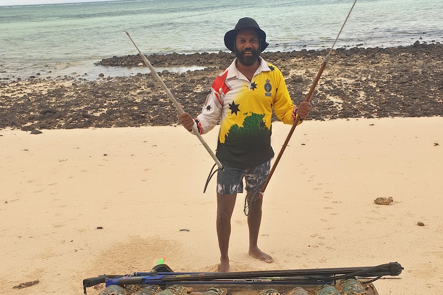 An Indigenous man on a beach holding a fishing spear in front of a table of crayfish.