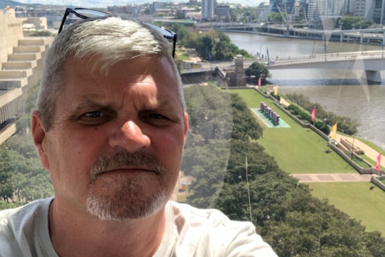 A man with grey hair and a beard takes a selfie from the Wheel of Brisbane above South Bank