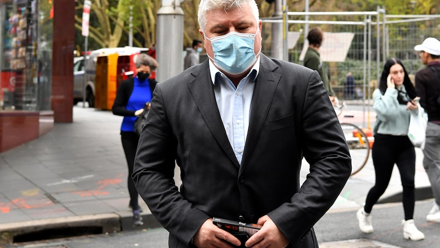 Stuart MacGill wearing a suit and a covid mask