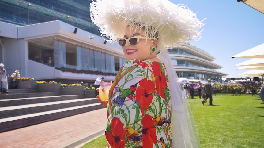 A woman wearing a red dress, white hat and sunglasses at the races.