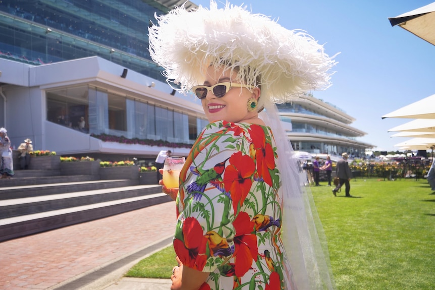 A woman wearing a red dress, white hat and sunglasses at the races.