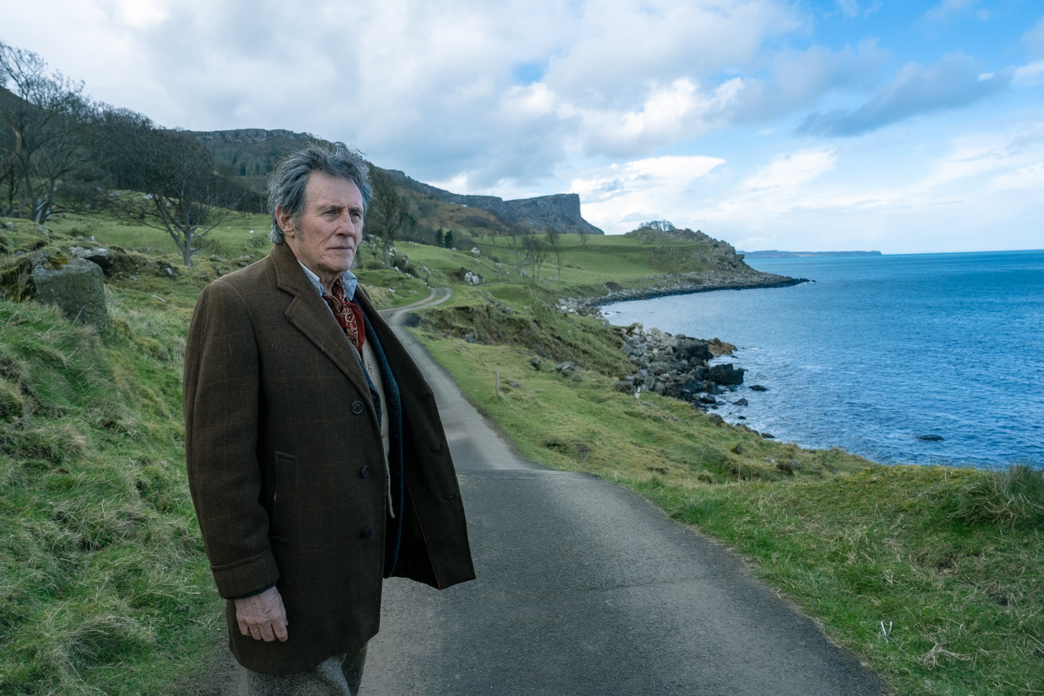 Gabriel looks out at sea while standing on a narrow bitumen road amid rolling green hills on a cold cloudy day.