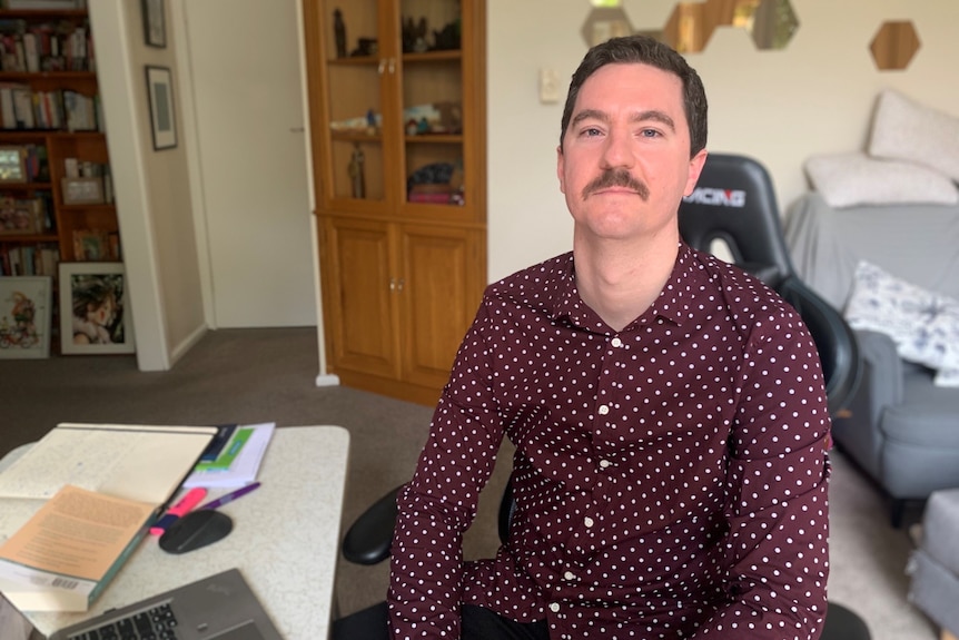 A young moustached man wearing polka dot burgundy shirt sits in front of laptop, looks at the camera.