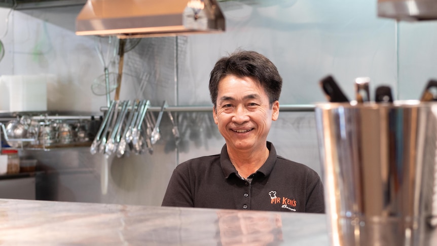 A middle-aged Cambodian man stands behind the pass of a commercial kitchen.