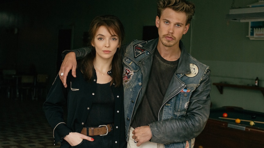 Jodie Comer and Austin Butler stand staring at the camera in 60s style clothes, his arm around her shoulders