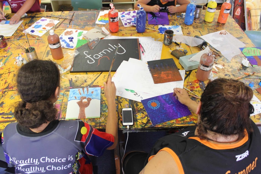 Aerial view of children sitting at a table covered in paint, colourful paper and paint brushes.