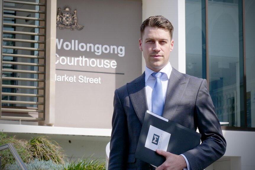 A man standing outside the Wollongong Courthouse wearing a suit holding a folder