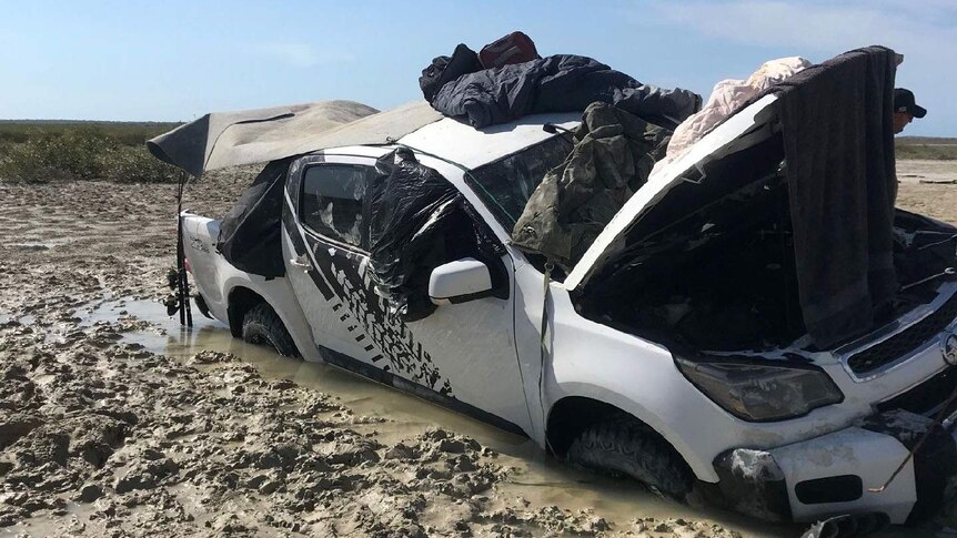 A four wheel drive heavily bogged in mud with its bonnet open and sleeping bags and gear on the roof.