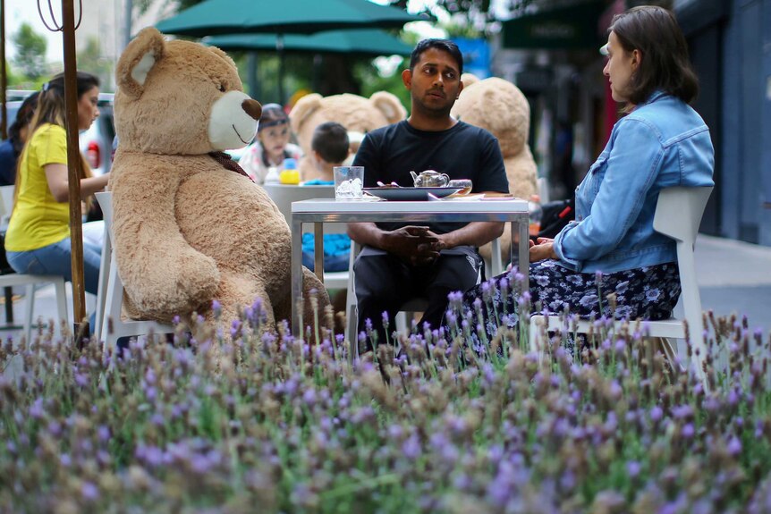 A man, a woman and a human-sized teddy bear sit at a cafe.