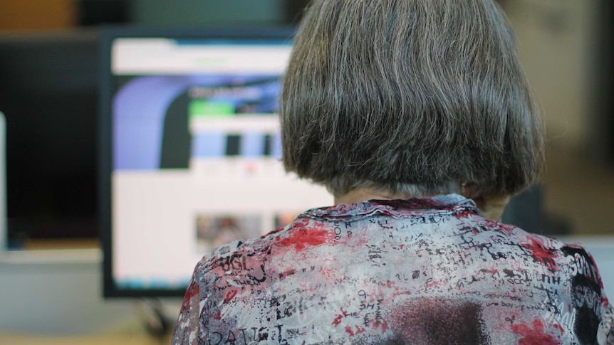 Back of grey-haired woman's head at desk with computer screen and take-away coffee cup