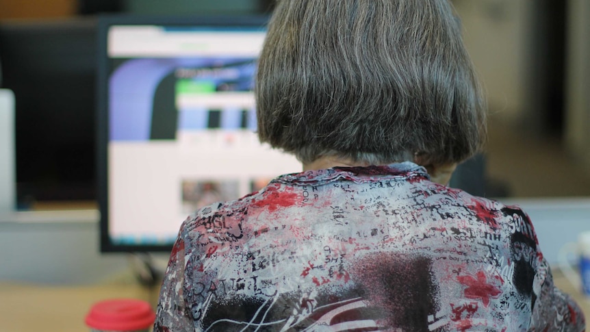 Back of grey-haired woman's head at desk with computer screen and take-away coffee cup