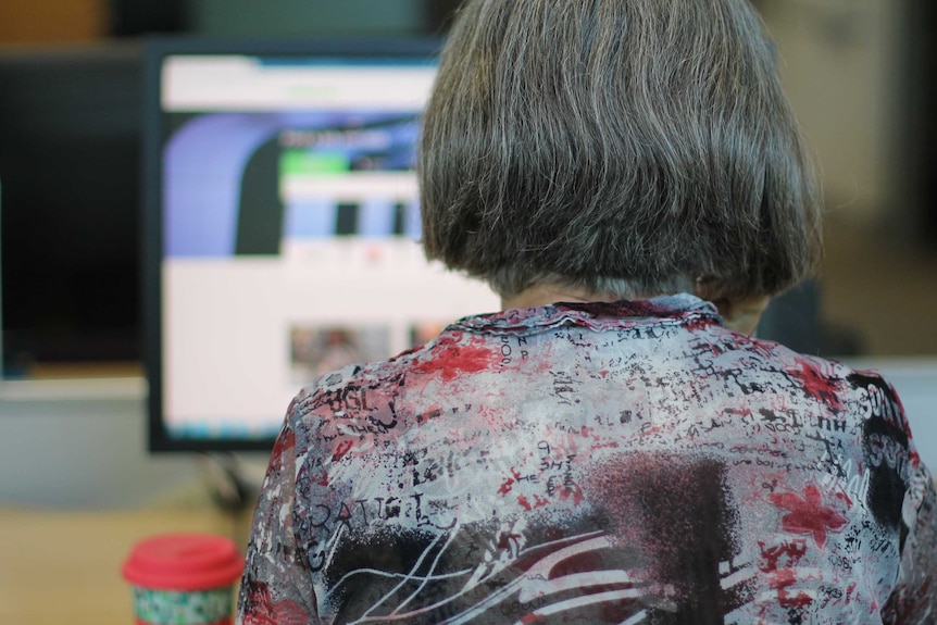 Back of grey-haired woman's head at desk with computer screen and take-away coffee cup