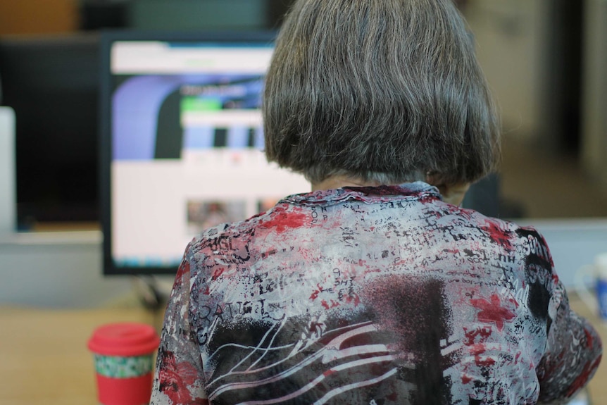 Back of grey-haired woman's head at desk with computer screen and take-away coffee cup