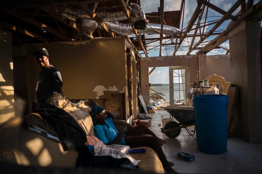 A man stands to the left while his daughter sits on a couch. Half the roof is missing off their home.