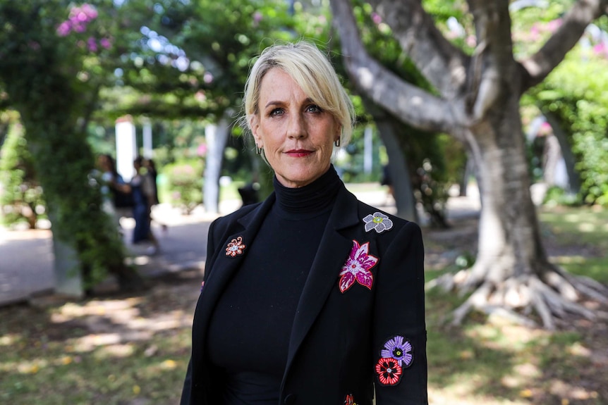 With blurred trees behind her, a woman with blonde hair; slight, closed-mouth smile; and black suit with floral design, stands.