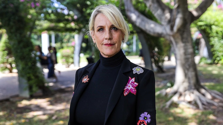 A woman with blonde hair wearing a dark suit with a floral design stands in front of some trees.