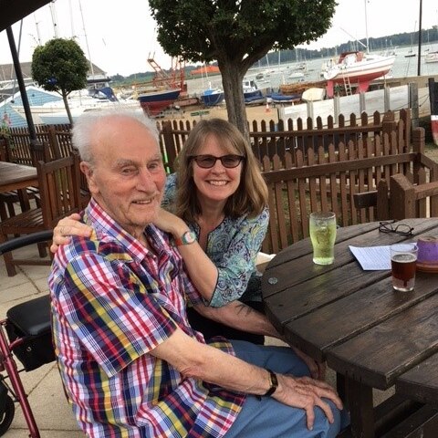 Sallie sits next to her father in a beer garden overlooking the water. There are multiple boats in the background.