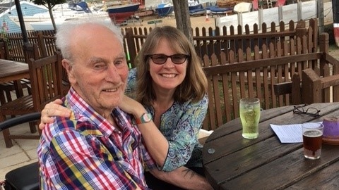 Sallie sits next to her father in a beer garden overlooking the water. There are multiple boats in the background.