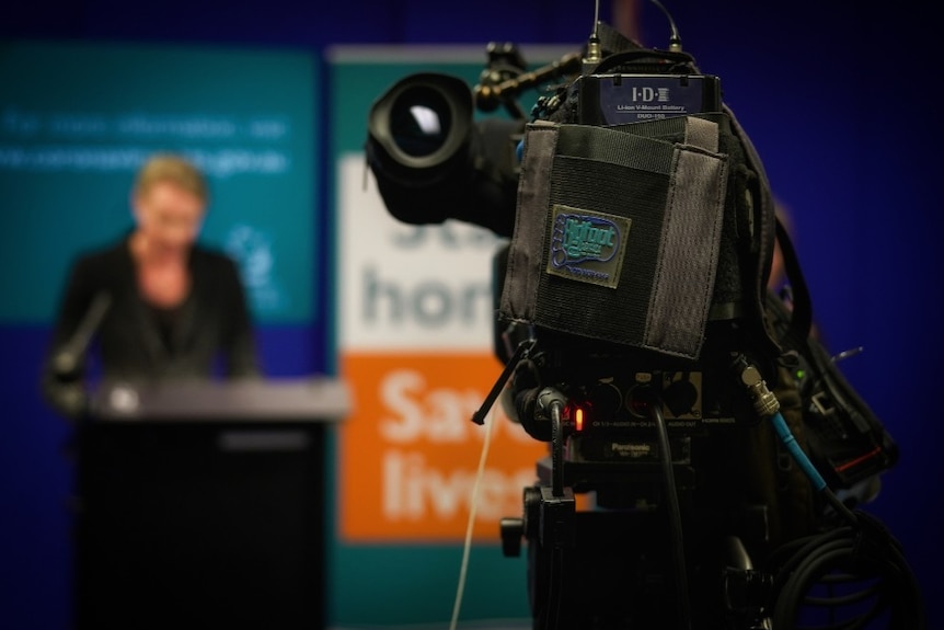 Camera at media conference with blurred person speaking at lectern.