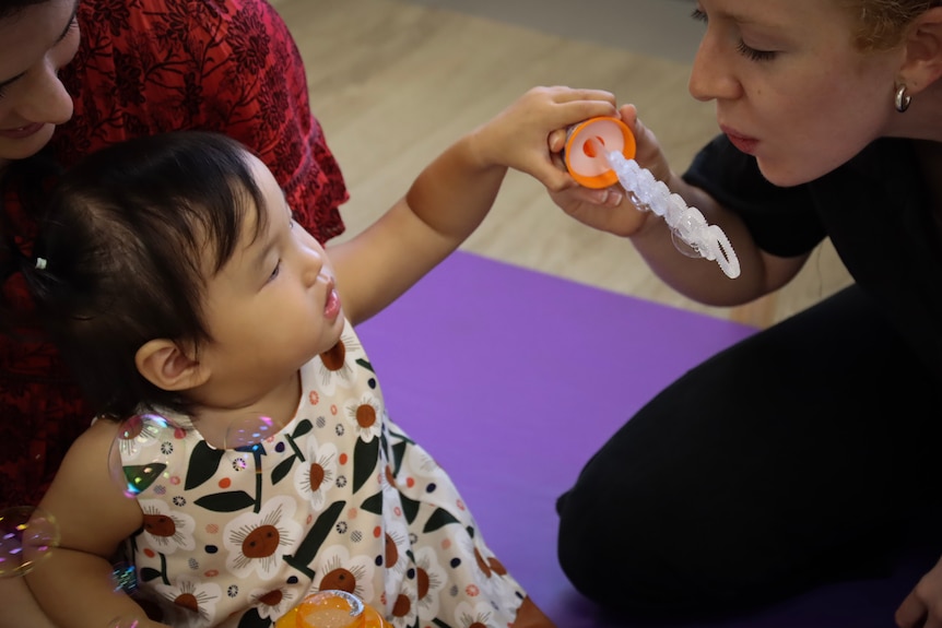 A woman wearing black pants and a shirt blows bubbles with a small girl. Everyone is happy.