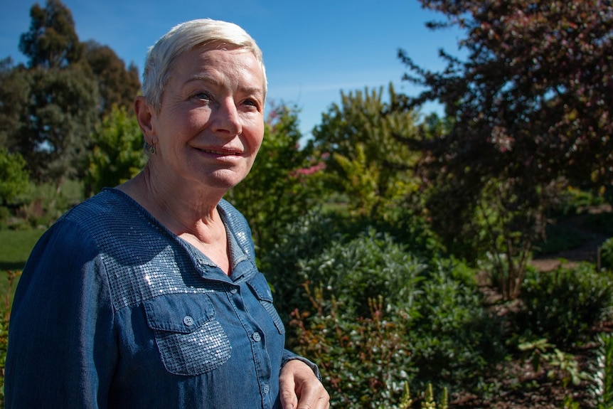 A woman with very short blonde hair wearing a blue denim shirt stares past the camera, surrounded by lush garden.