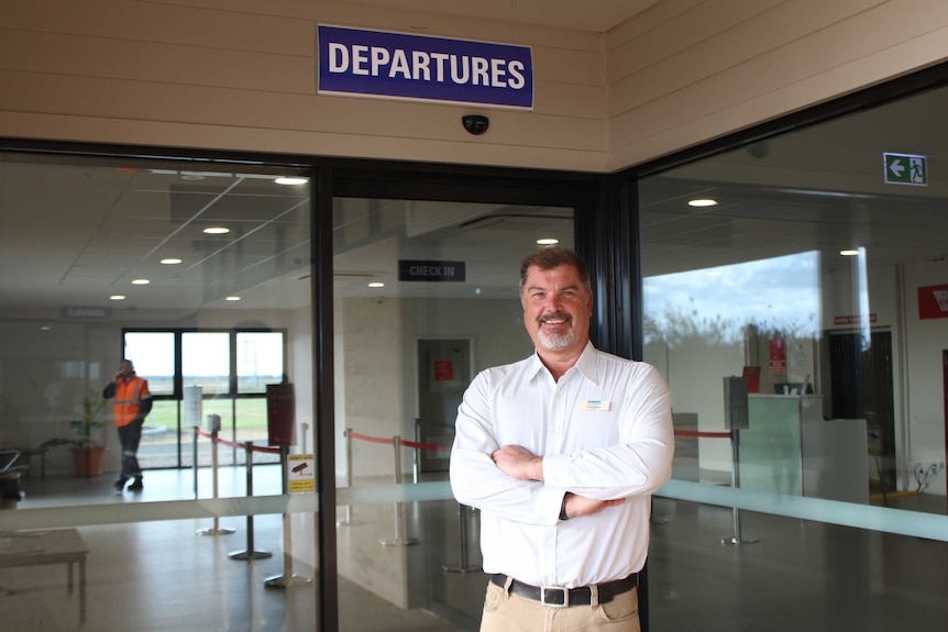 Busselton Mayor Grant Henley stands with his arms folded inside the town's airport terminal, under a departures sign.