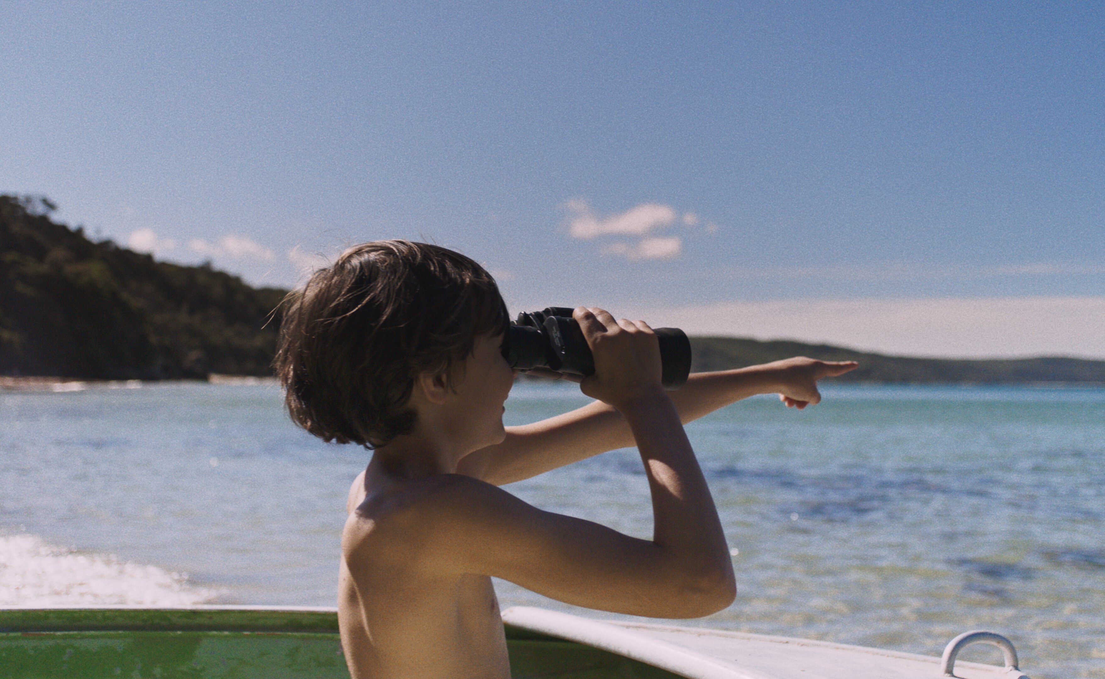 A young boy holds binoculars in one hand and points with his other hand. Clear ocean water surrounds him. 
