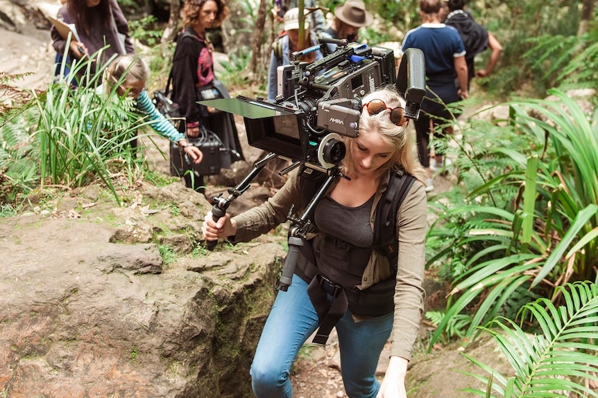 Film crew carrying camera climbing up rocky trail rainforest