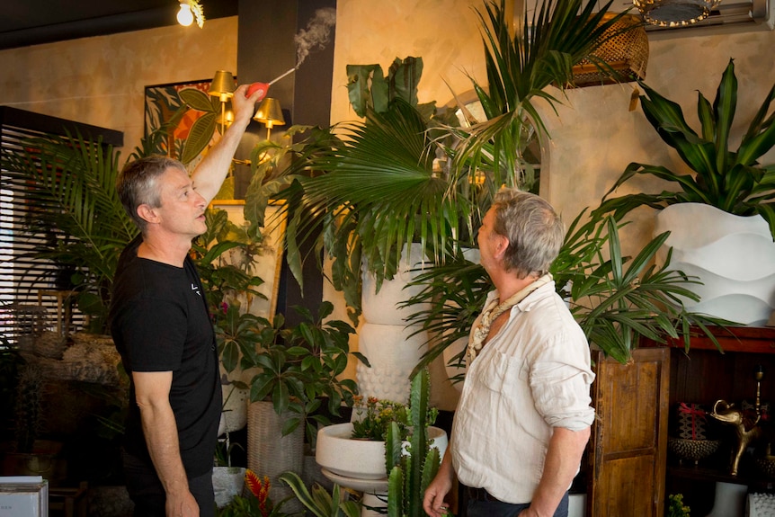 Andrew Orfanos holds a smoke tube up near an airconditioning system while the owner watches the process.