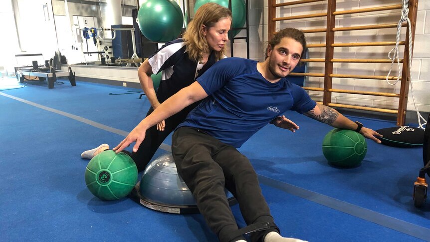 Sam Tait leans to his left with both arms outstretched, balancing on medicine balls, Kelly Beahan crouches behind him