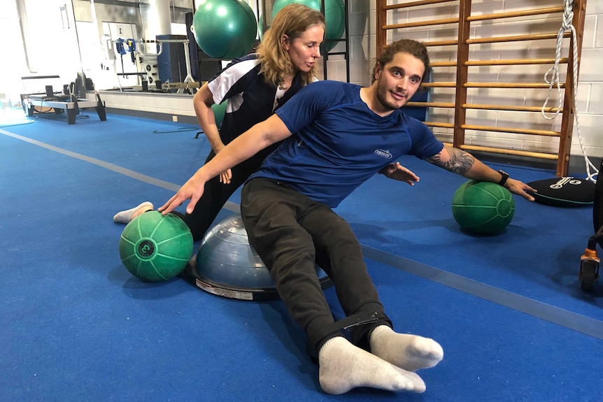 Sam Tait leans to his left with both arms outstretched, balancing on medicine balls, Kelly Beahan crouches behind him