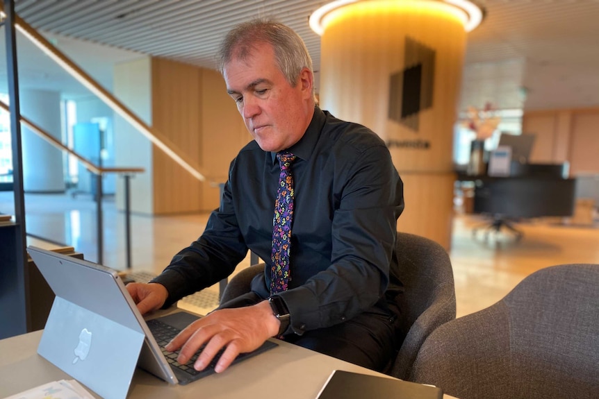 A man wearing a black shirt and tie sits at a desk working on his laptop.