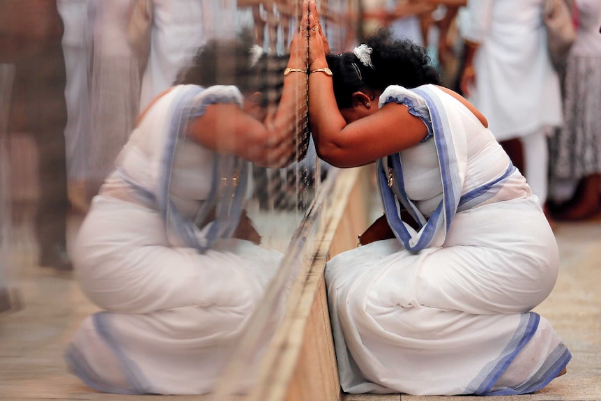 A woman in a white and blue dress presses her hands up against a reflective black commemorative wall with her head down.