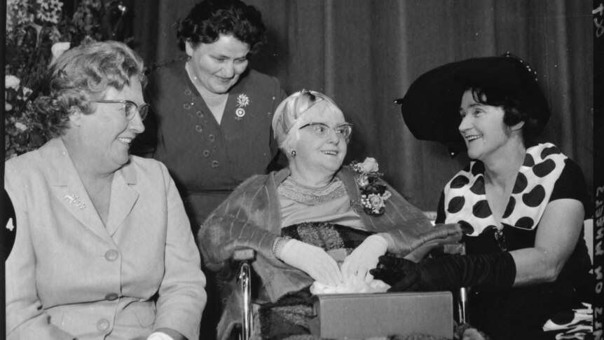A black and white photograph of four older women, smiling at a formal occasion. Doris Taylor is seated in her wheelchair. 