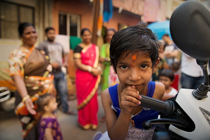 A street shot from the Kanjabhat community in Pune. A girl is standing holding onto a motorbike handle.