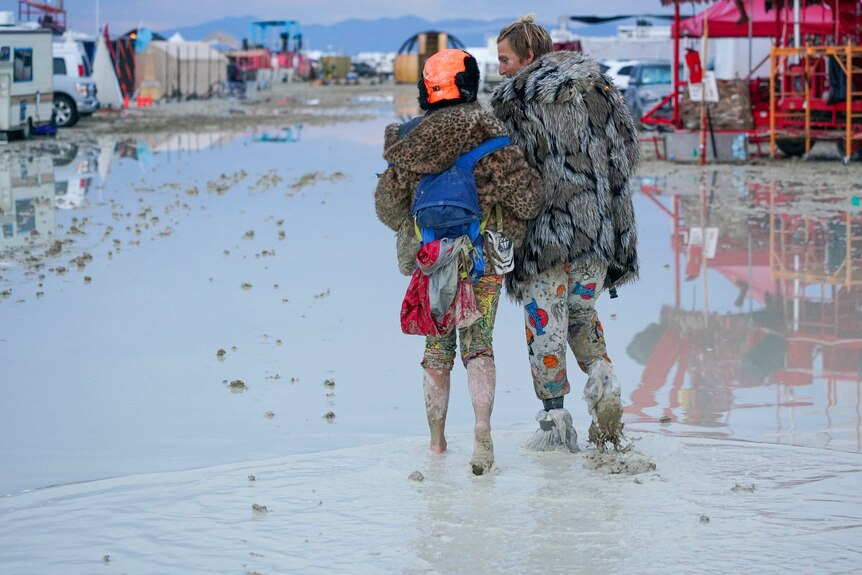 Two people wearing colourful festival clothes walk through mud with their backs to the camera.