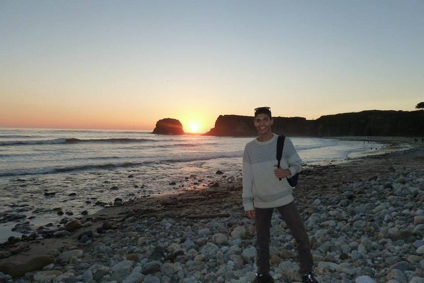 Writer and comedian AJ Lamarque wears a jumper and holds a backpack while posing on a beach as the sun goes down.