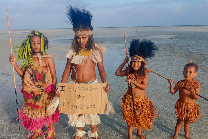 Four Torres Strait Islander childen pose on the beach, the tallest holding a sign in support of the Torres Straight 8.