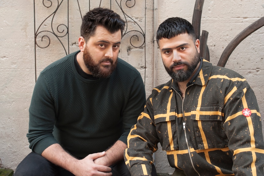 Two men sit in front of an ornate gate. They both have serious expressions, short beards and short hair 