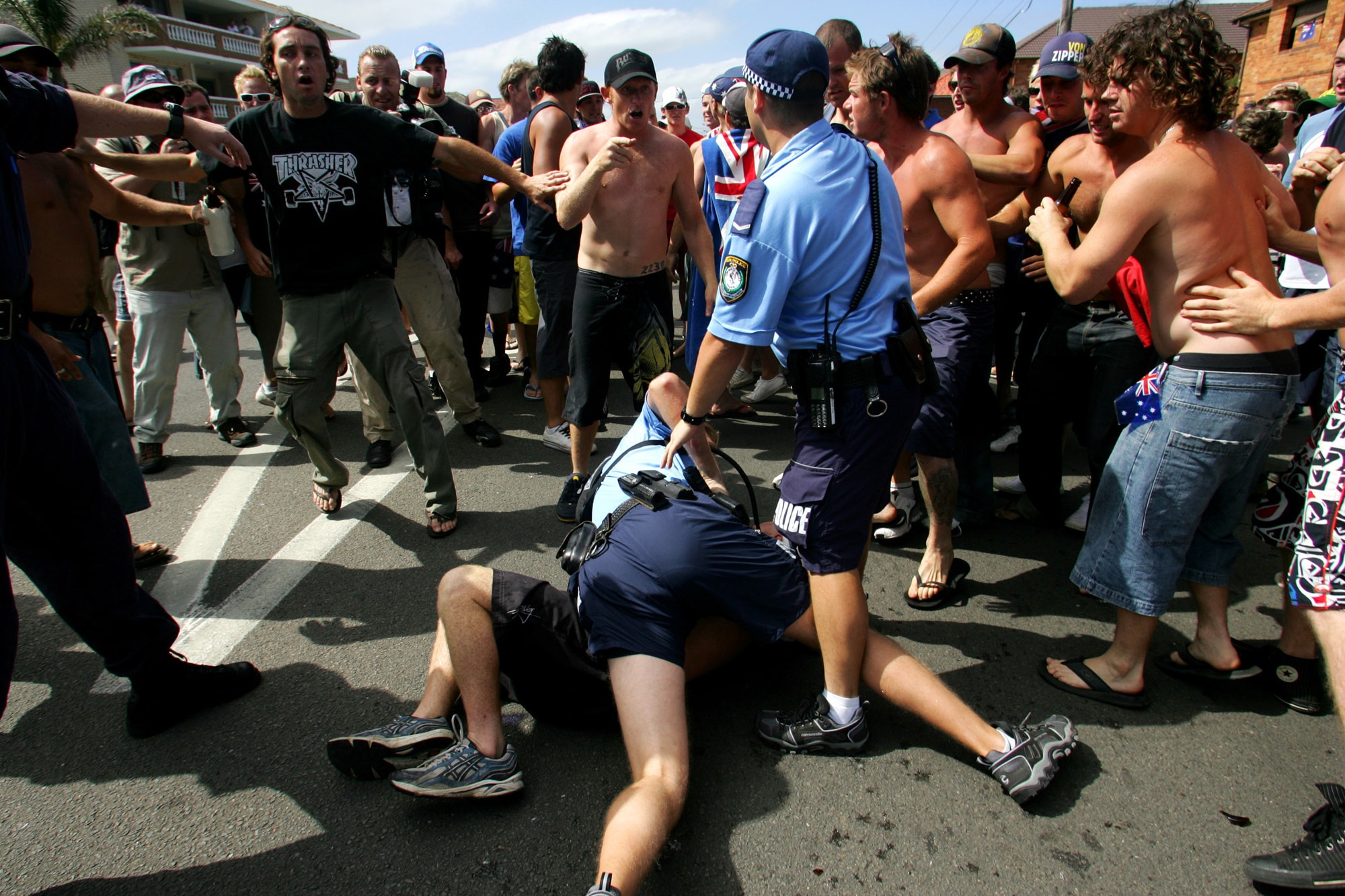 Two police officers arrest a person on the ground as shirtless men surround them.
