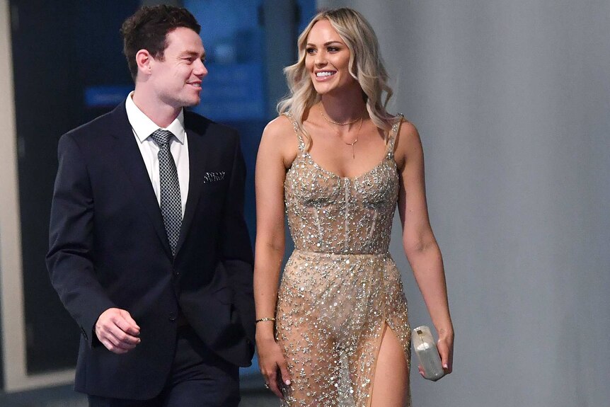 Lachie Neale wears a suit as he walks next to Julie Neale, who wears an embezzled sheer gown and wears her hair in waves.