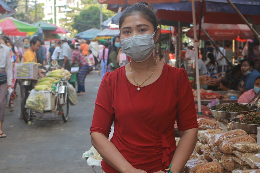 A woman wearing a red shirt and a mask with a ponytail stands in a market with umbrellas.