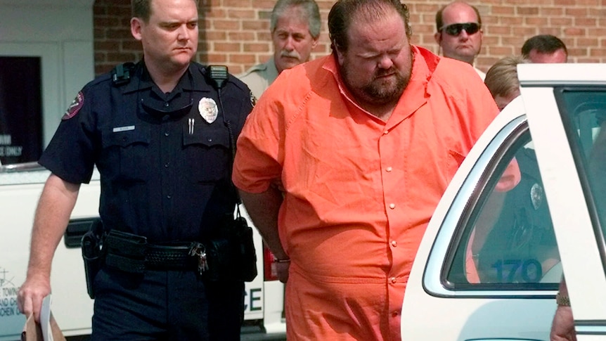 A large man in an orange jumpsuit is led to a car by a police officer. 