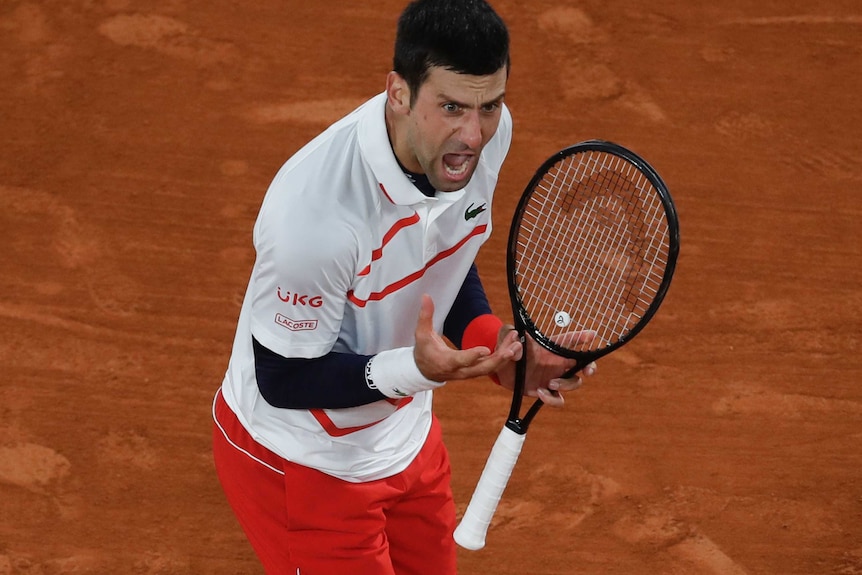 Novak Djokovic gestures with his hand and yells in frustration while holding a racquet. He is standing on a clay court