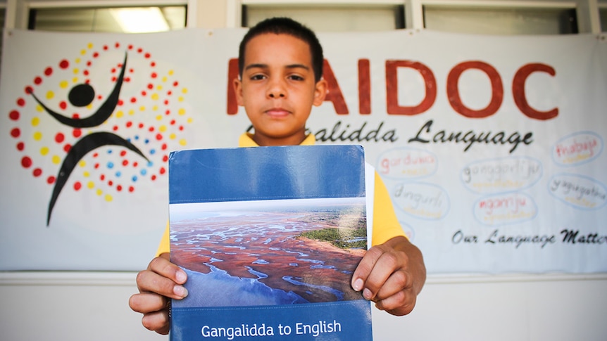 Tjabadungah Yanner holding the Gangalidda to English dictionary, standing in front of a NAIDOC sign.